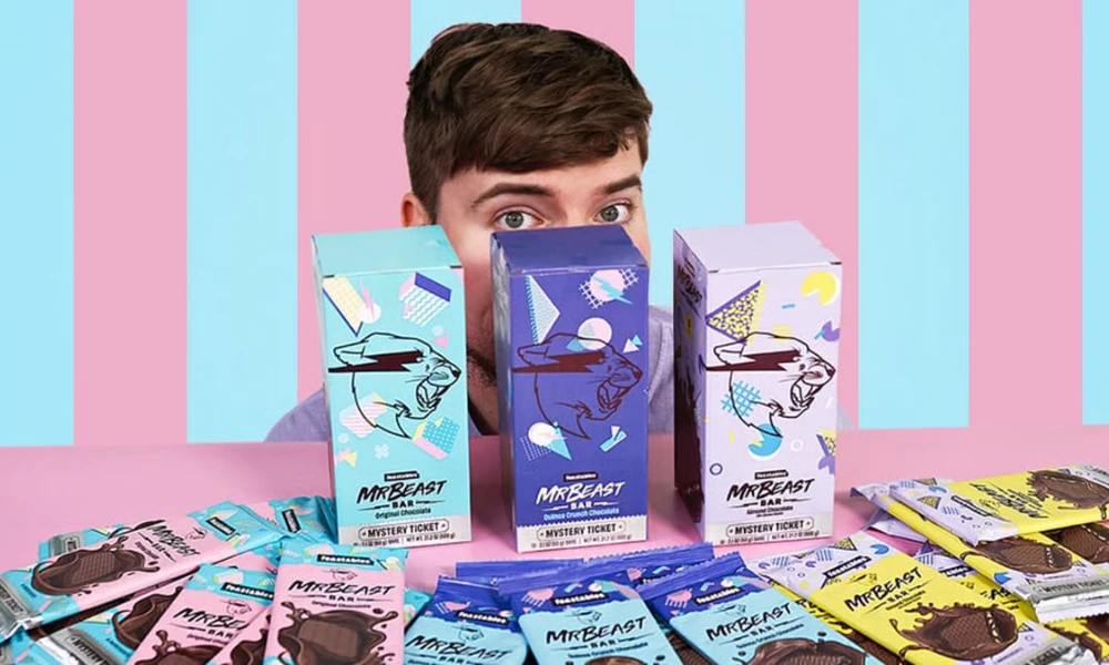| Feastables: MrBeast's $250M Chocolate Empire & Why It's More Profitable Than YouTube Three custom Feastables merchant display boxes standing on a counter desk against a striped backdrop