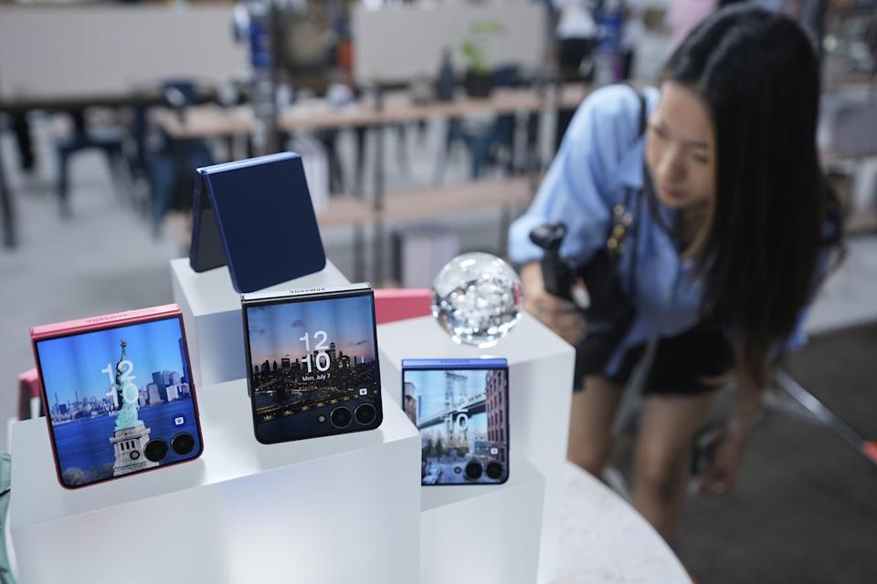 | Samsung Galaxy Fold 8 Leaks Reveal Major Design Changes A retail experience zone display counter showing four pastel pink and blue clamshell flip phones with a blurry consumer reviewing tech products in the background