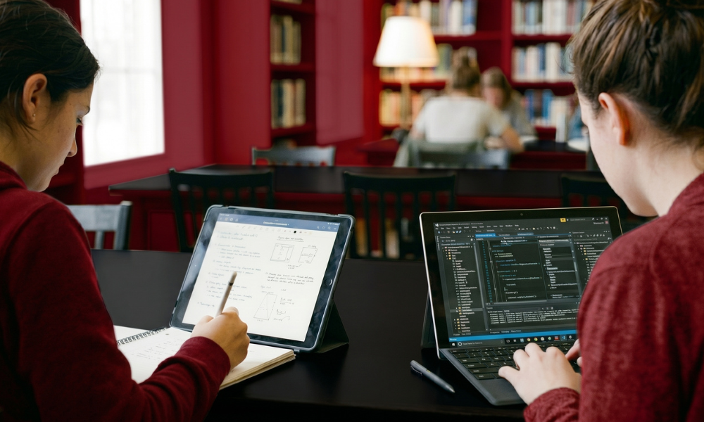 | iPad vs Microsoft Surface: Best Tablet for Productivity in 2026? Students using digital stylus note taking software and an advanced code programming IDE terminal in a university library