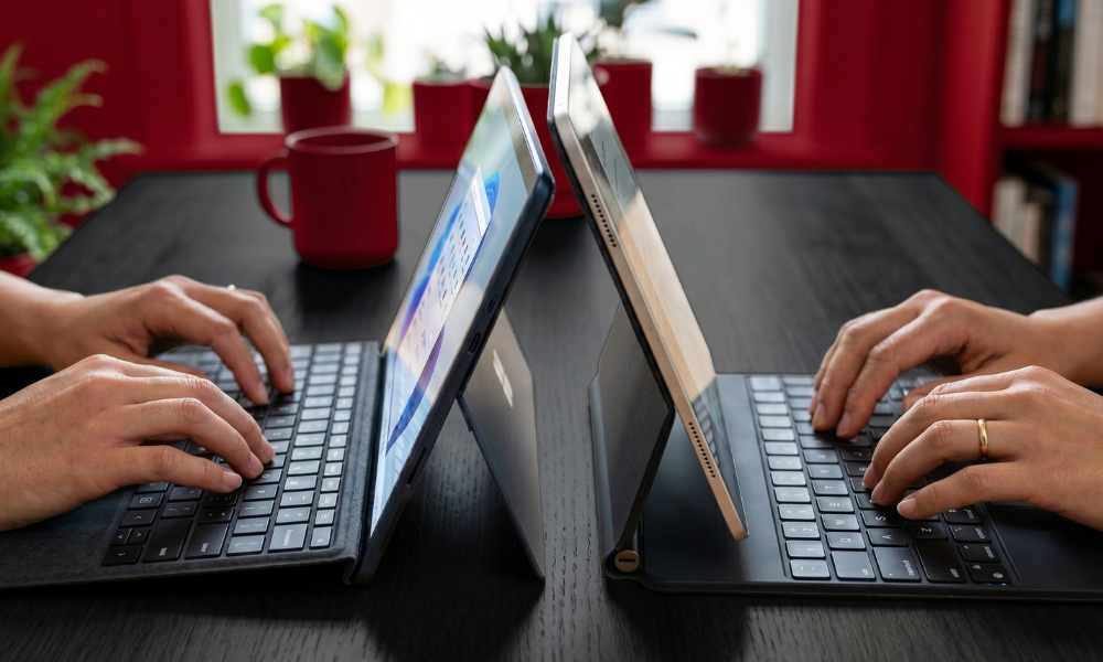 | iPad vs Microsoft Surface: Best Tablet for Productivity in 2026? Two users concurrently typing on a Microsoft Surface detachable keyboard case and an Apple iPad Magic Keyboard folio