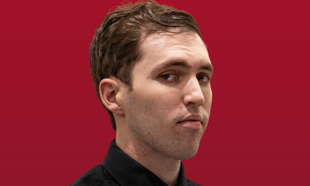 | Tyler Robinson Case: How Political Violence Is Changing America A close-up mugshot profile of Tyler James Robinson against a solid red studio backdrop portrait setting