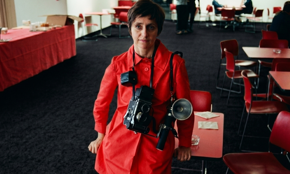 | Diane Arbus: The Complete Guide to America's Most Important Photographer of the Margins (2026) A full length portrait of an artist in a red coat with a twin lens Rolleiflex camera and flash unit hanging from her neck