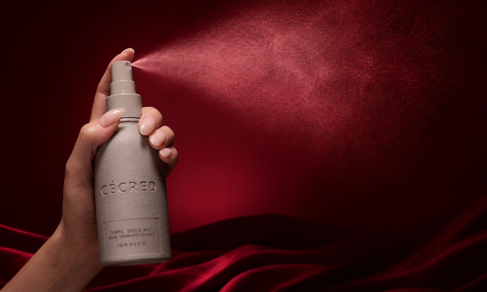 | Cécred Hair Products Review: Beyoncé's Complete 2026 Guide A hand spraying a fine cloud mist from a grey canister of Cécred Thermal Shield Mist against a rich crimson ambient background