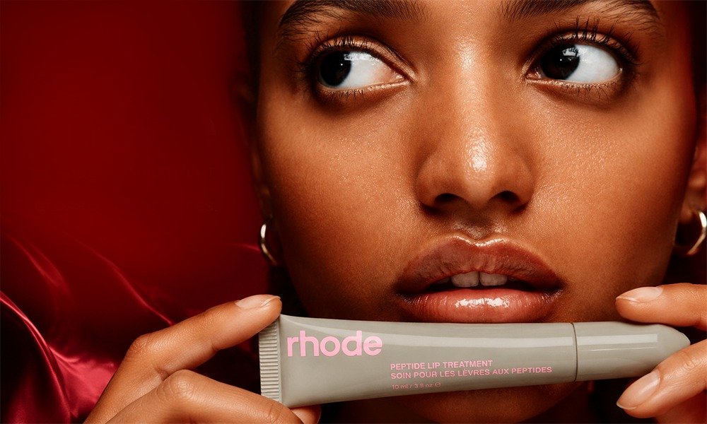 | Rhode Lip Peptide Treatment Review: Flavors & Buying Guide (2026) A macro detail portrait of glossy lips close to a gray tube of Rhode Peptide Lip Treatment with pink lettering