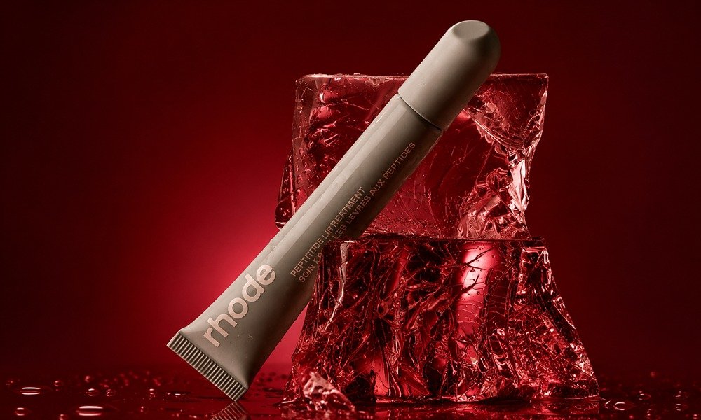 | Rhode Lip Peptide Treatment Review: Flavors & Buying Guide (2026) A gray tube of Rhode Peptide Lip Treatment resting diagonally on a jagged red crystalline block of crushed ice