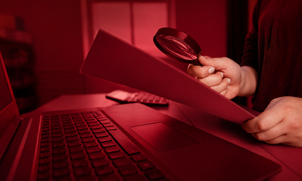 Person using a magnifying glass to review a document near a laptop.
