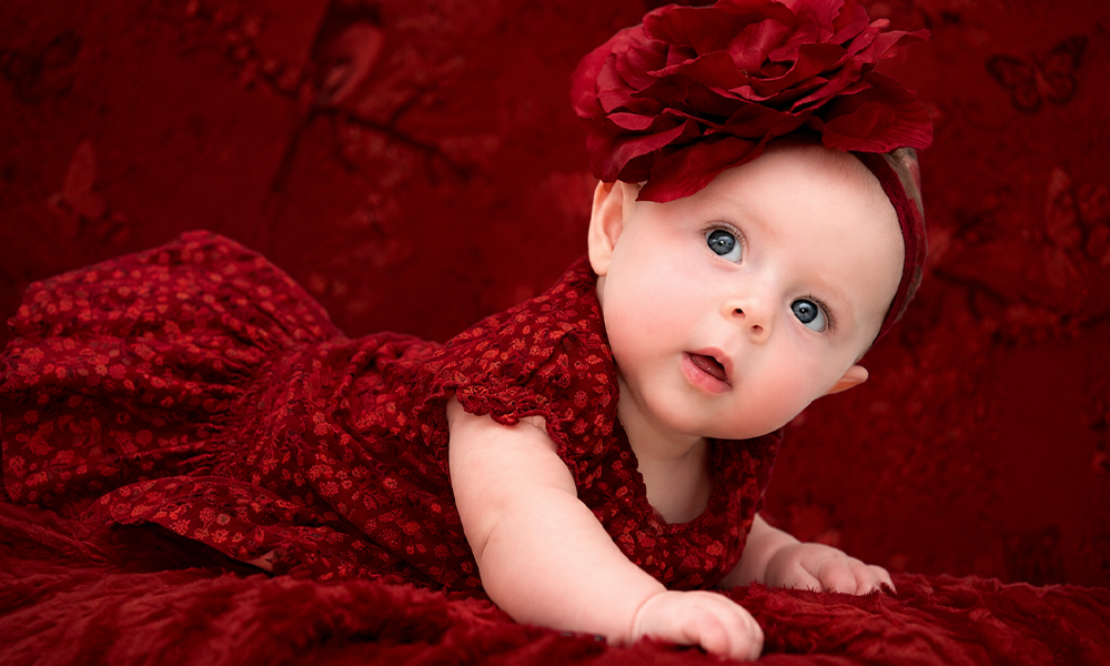 Baby in a red floral dress and flower headband lying on a red blanket.