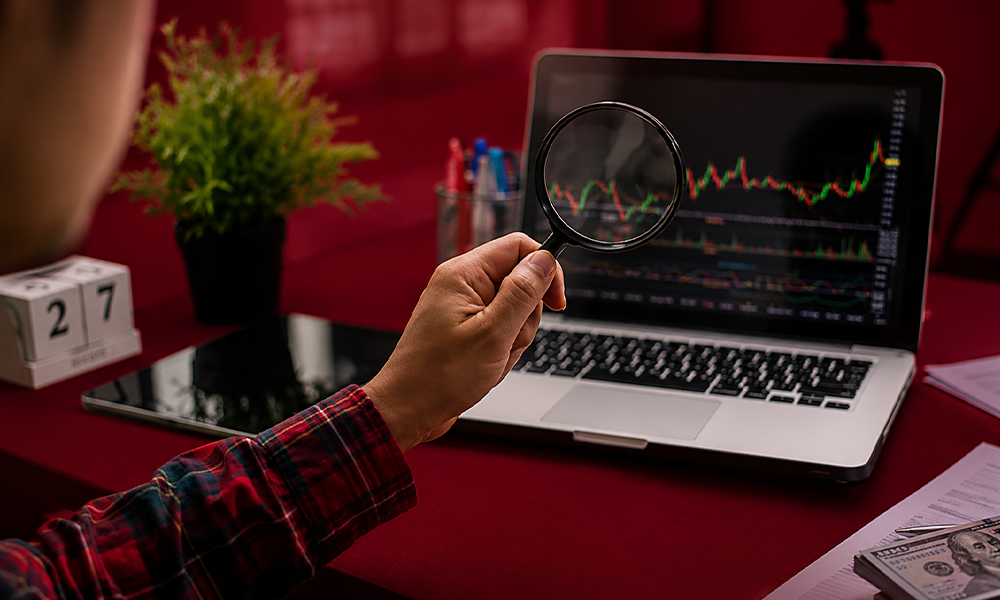 Magnifying glass examining stock market data charts on a laptop screen.