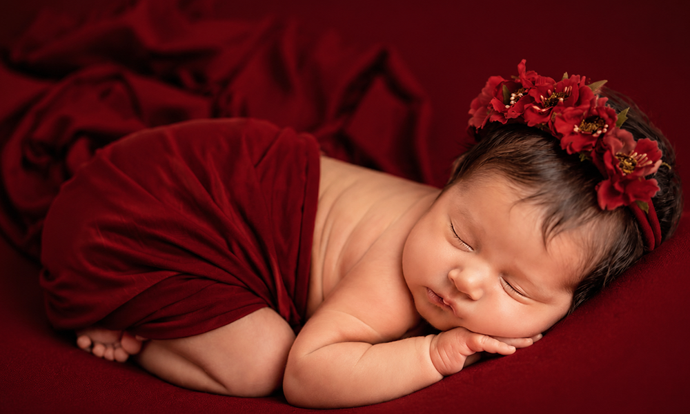 Sleeping newborn baby wrapped in red fabric wearing a floral wreath.