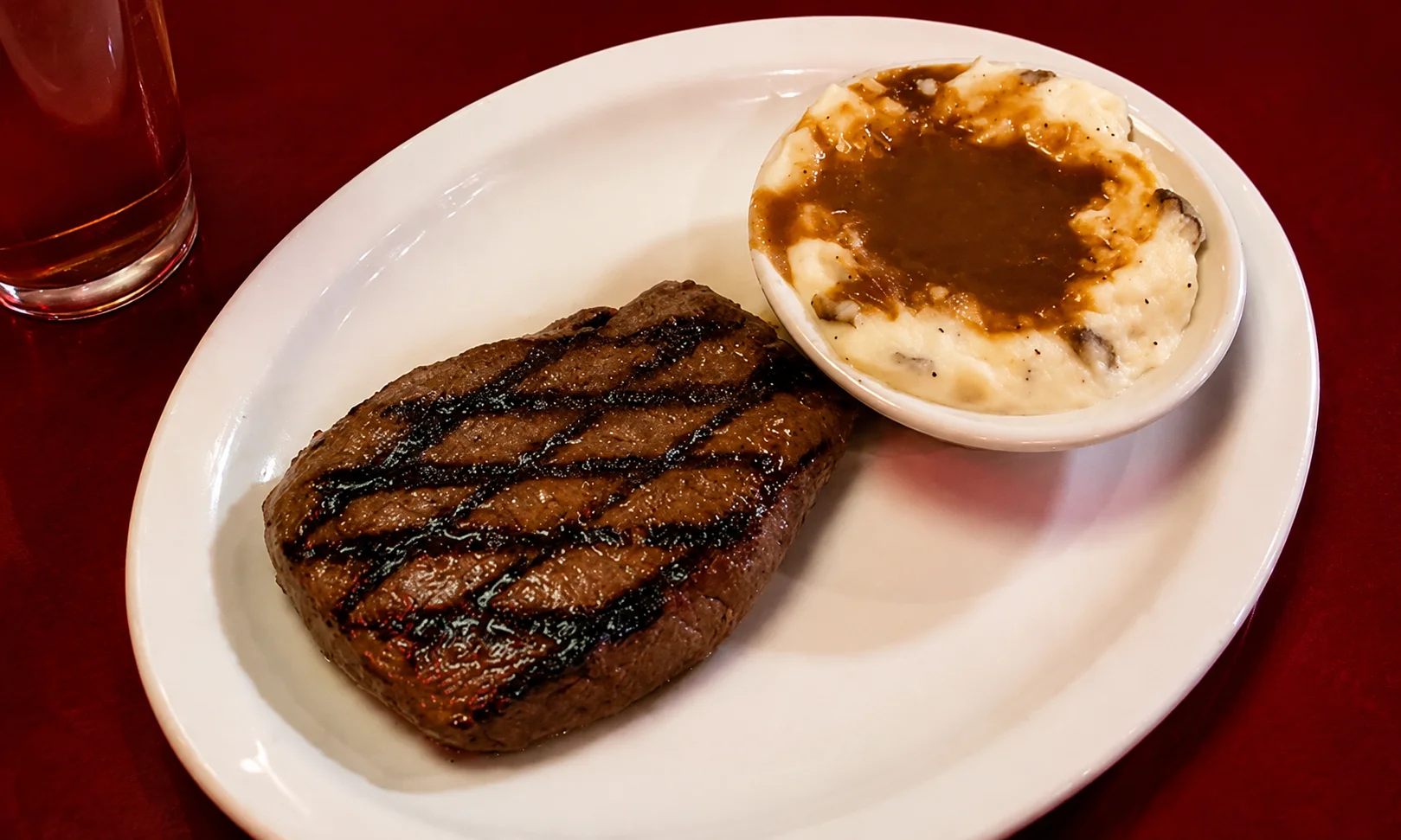 A grilled steak on a white plate with a side dish in a red-lit dining setting