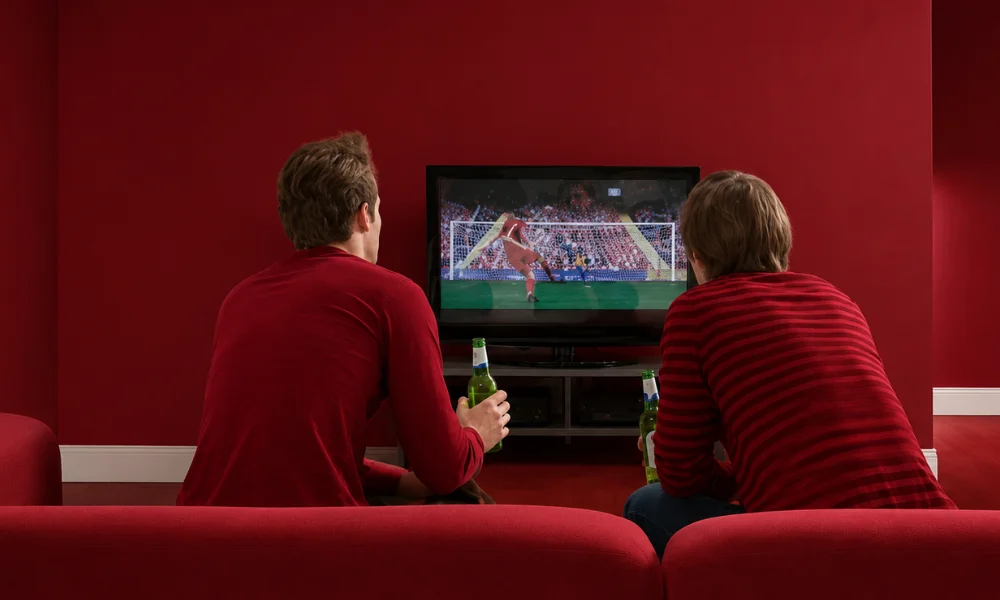 Two people sitting on a red sofa, watching a live sports broadcast on a television in a red-themed room.