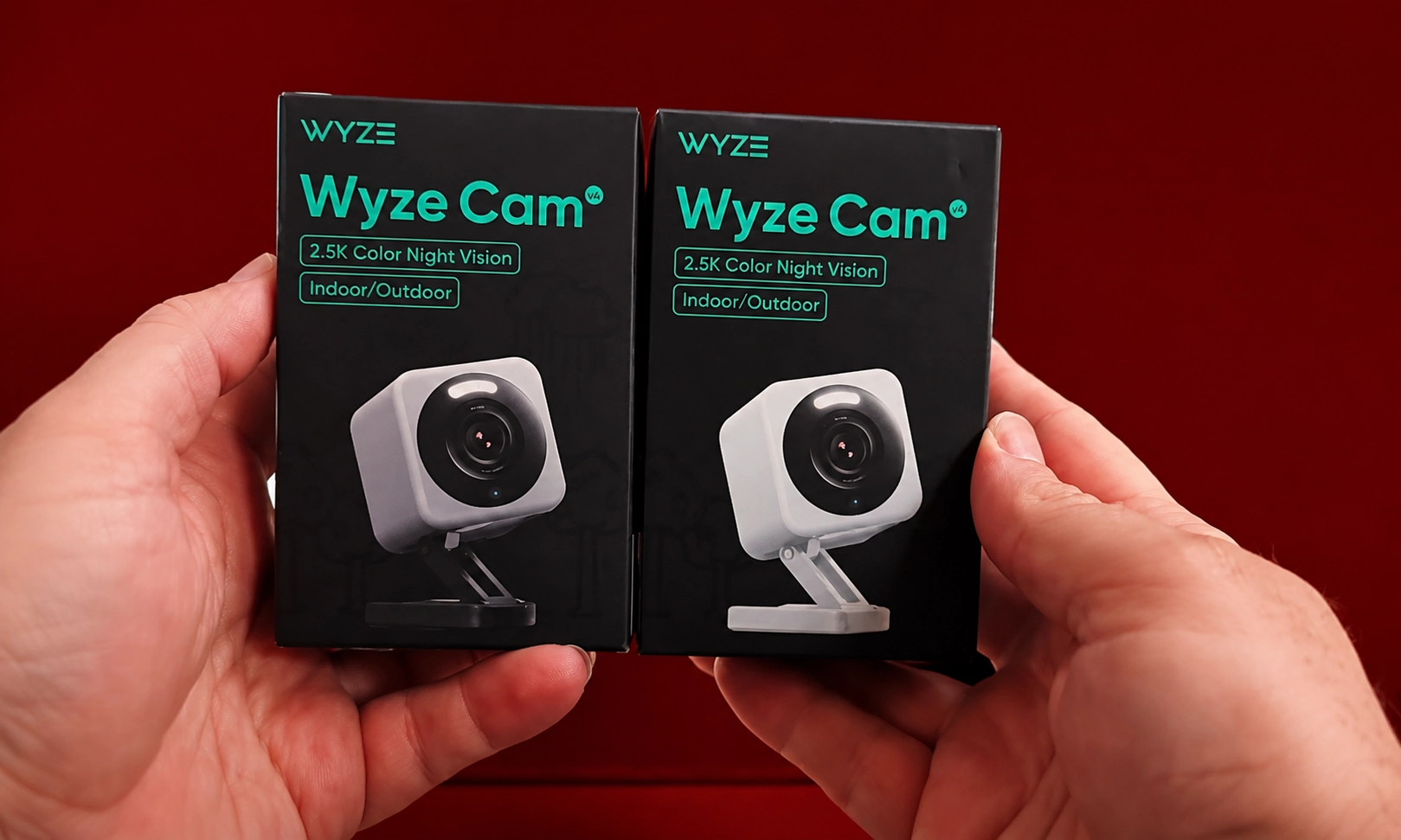 A pair of hands holding security camera packaging or small devices against a dark red background.