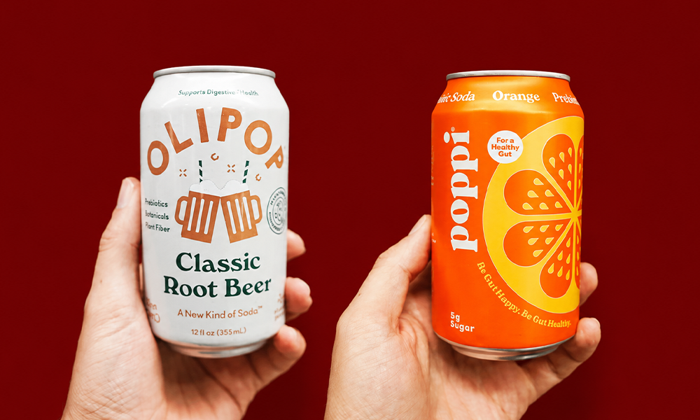 | Poppi vs Olipop: A Complete Side-by-Side Comparison (2026) view of hands showcasing a can of Olipop Classic Root Beer and an orange Poppi soda against a solid deep red background