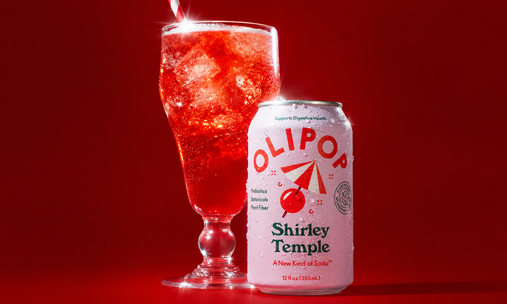 | Poppi vs Olipop: A Complete Side-by-Side Comparison (2026) A pink can of Olipop Shirley Temple next to a sparkling glass filled with iced red soda and a striped straw