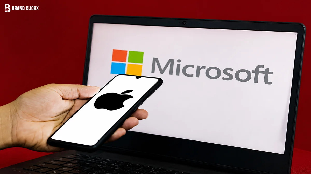 Person holding a smartphone displaying an Apple logo in front of a laptop showing the Microsoft logo