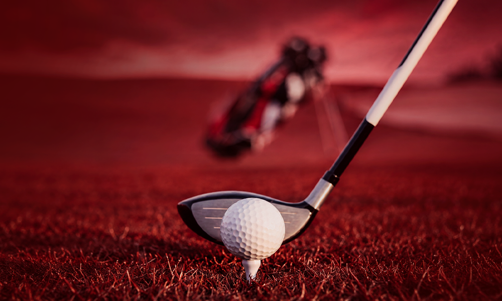 A golf club swinging near a golf ball positioned on a red-toned turf or course surface.