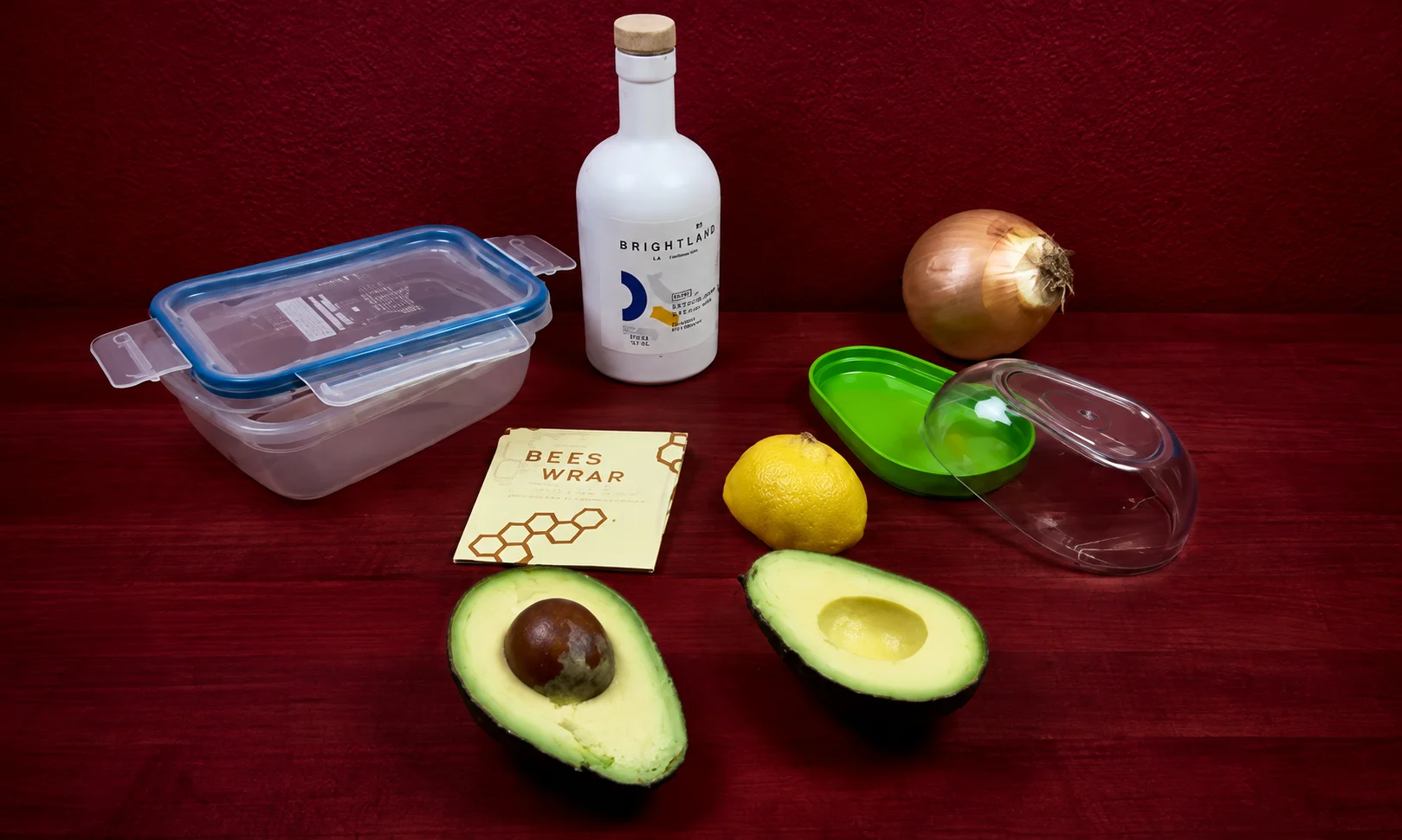 Assorted storage containers and halved avocados arranged on a dark red surface.