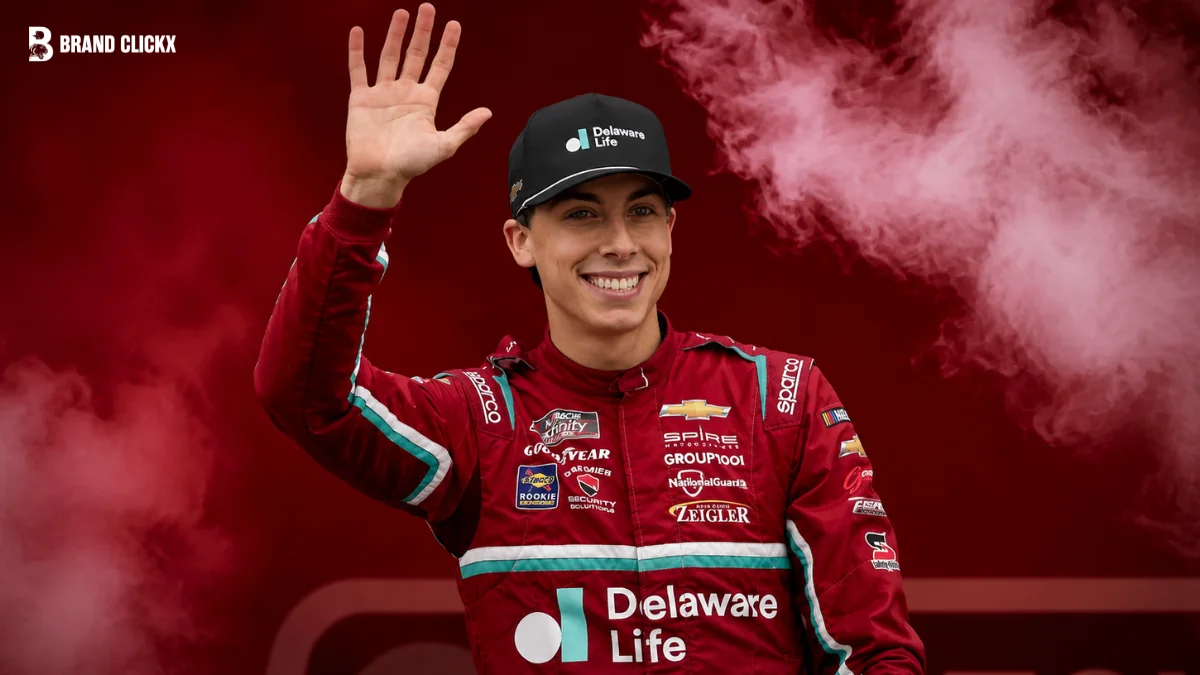 Portrait of Carson Hocevar waving in racing gear set against a dark red ambient background.