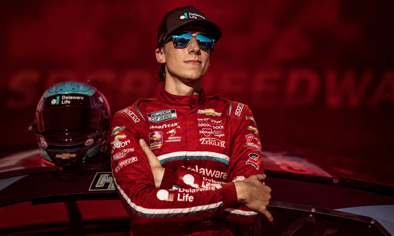 Portrait of Carson Hocevar in a red racing suit displayed against a rich red studio backdrop.