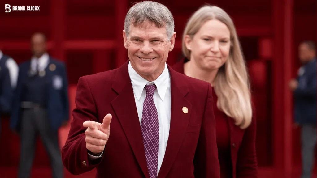 A smiling man in a red blazer pointing forward against a dark red background