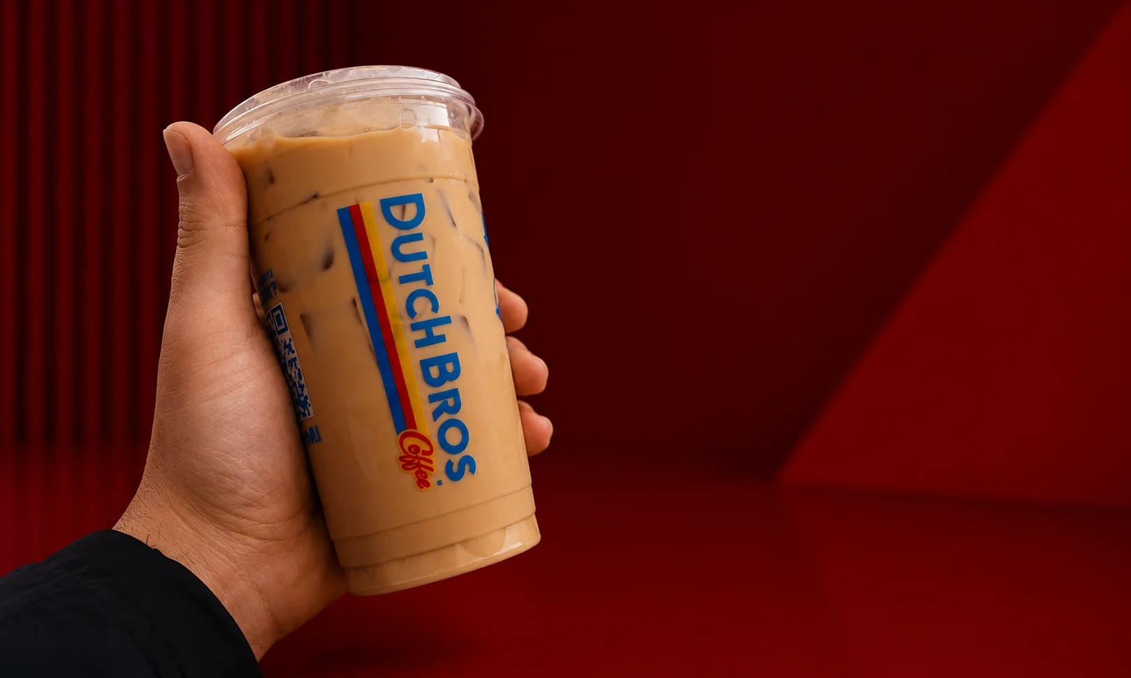 A hand holding a Dutch Bros cup against a rich red background.
