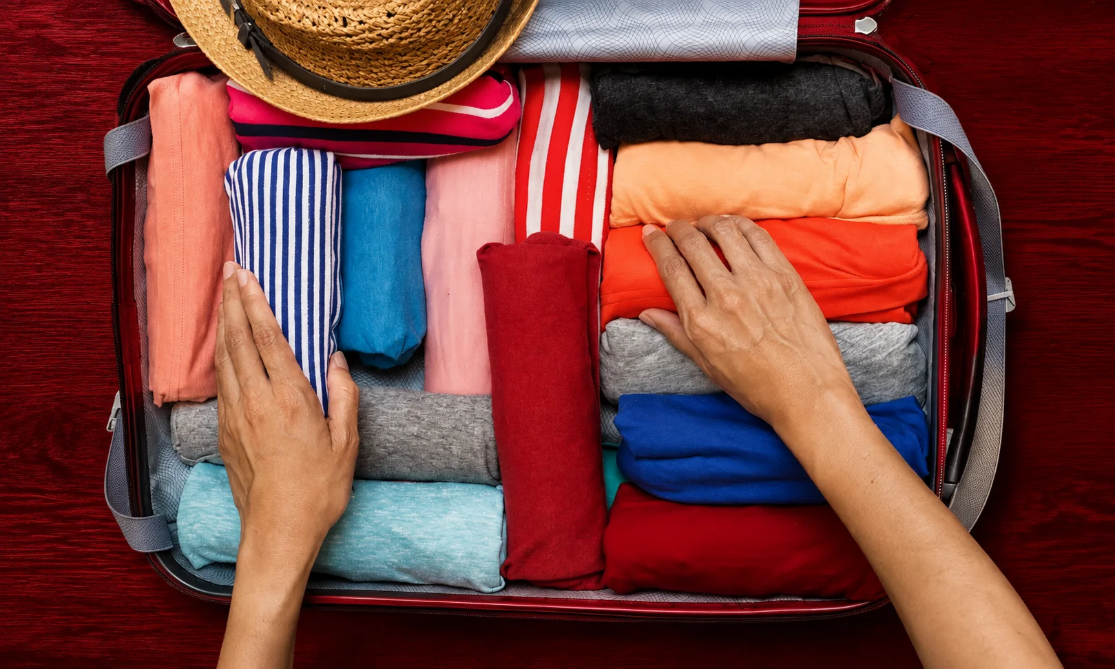 Hands organizing packing cubes and clothes inside an open suitcase against a dark red 