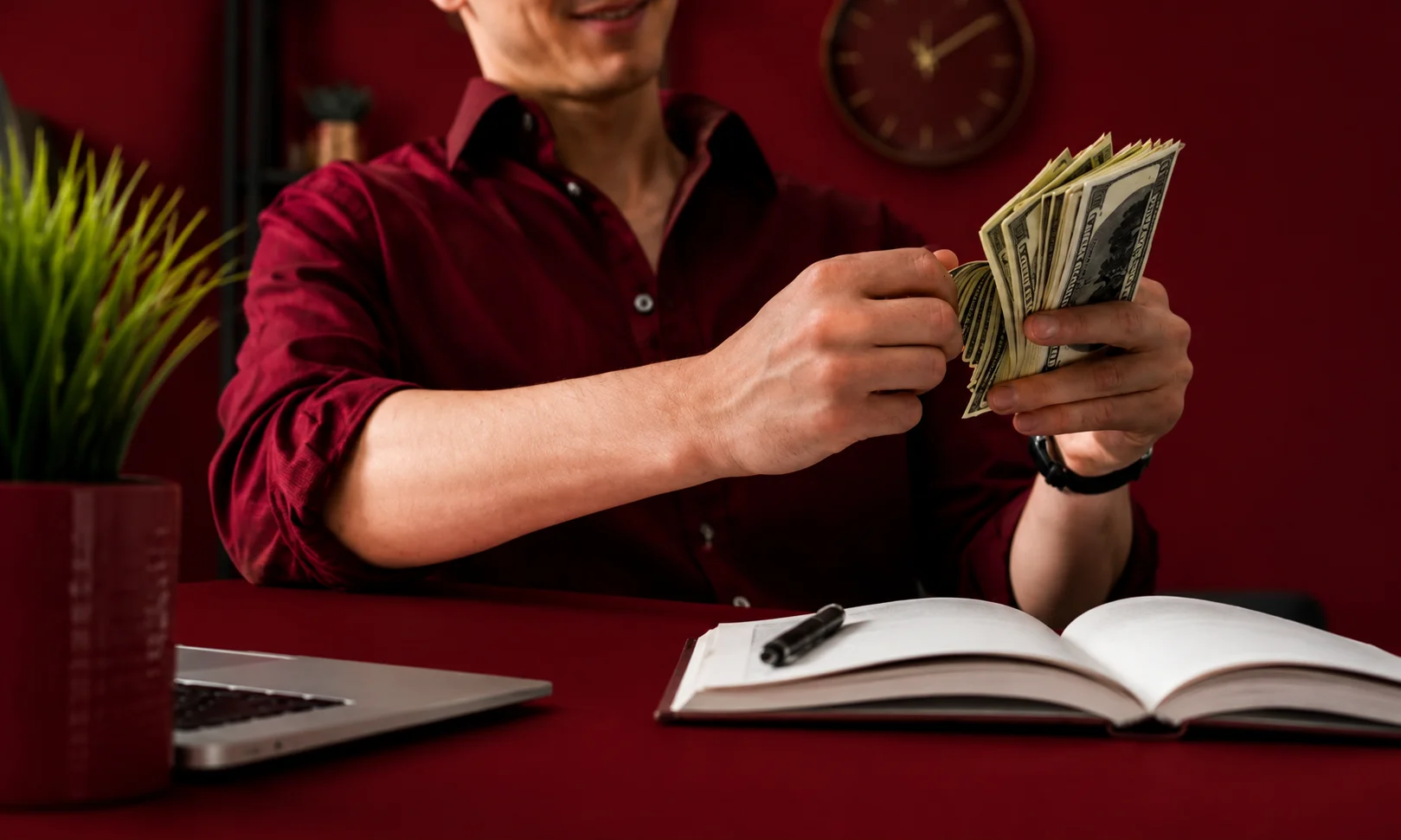 A person holding a stack of cash at a desk with a notebook, set against a dark red background.