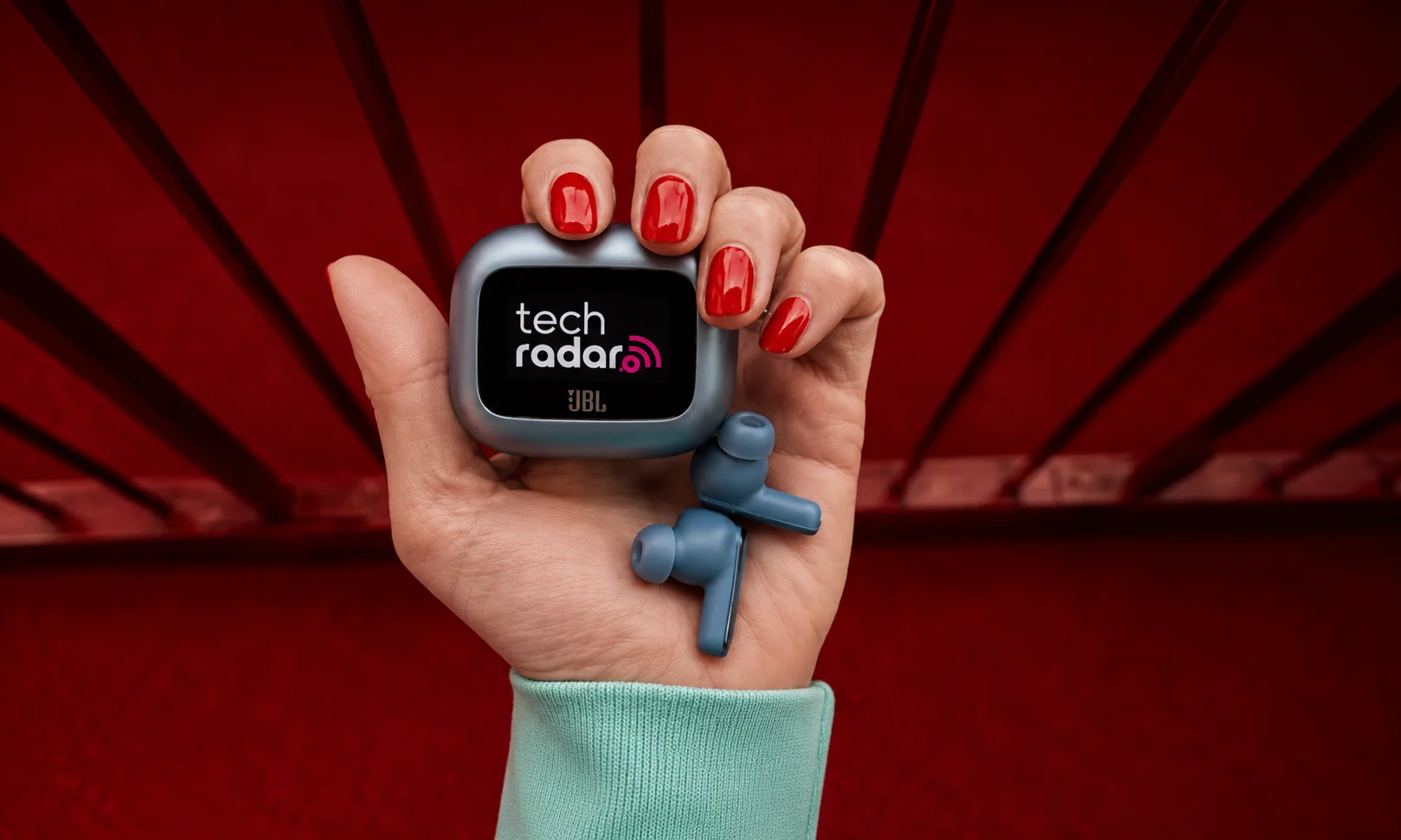 A hand holding JBL earbuds against a red studio backdrop.