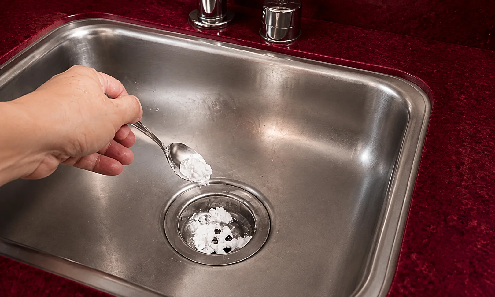 A hand applying baking soda into a stainless steel sink set against a dark red ambient background.