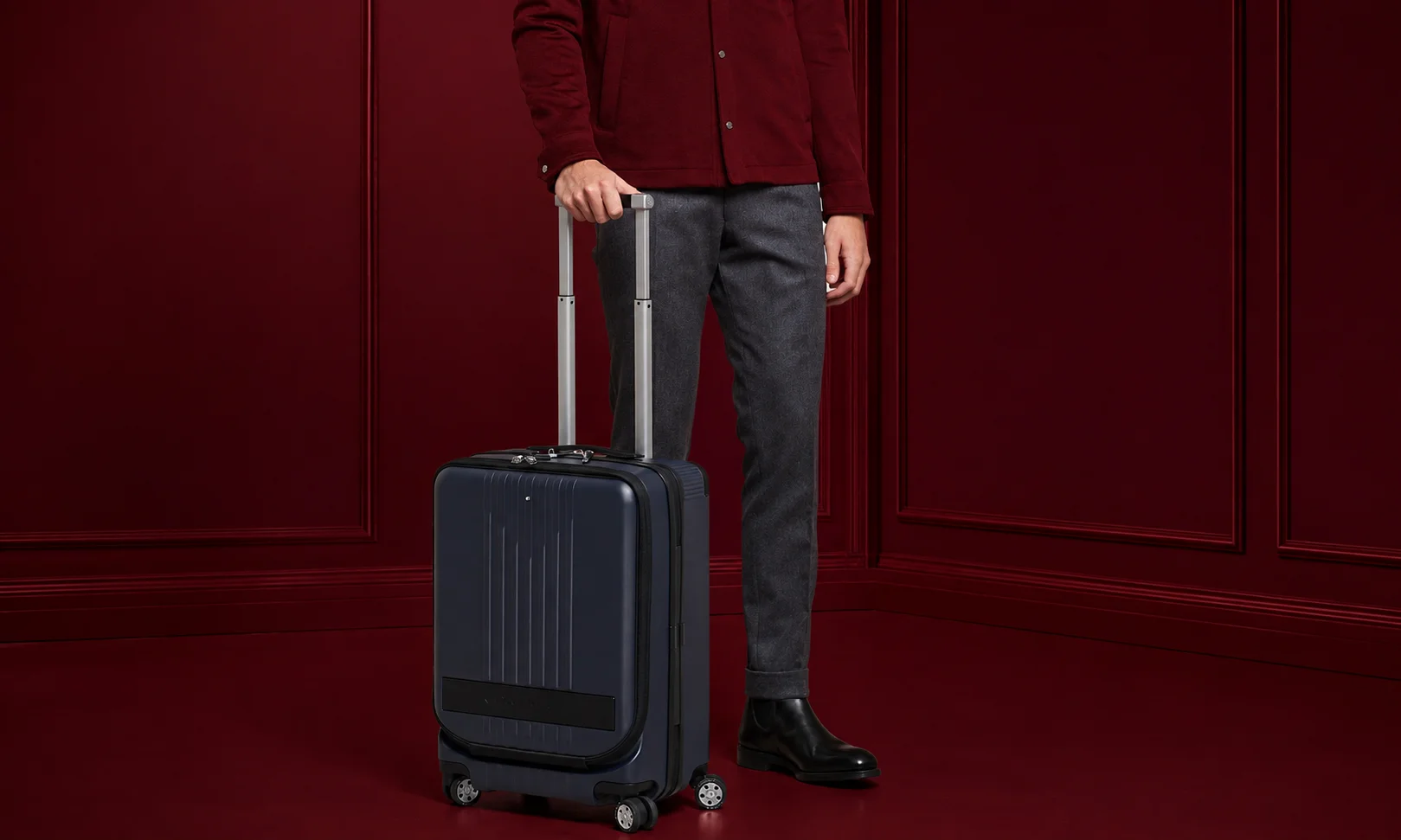 | 15 Best Luxury Luggage Brands for Travelers in 2026 A traveler with Montblanc luggage against a dark red backdrop.