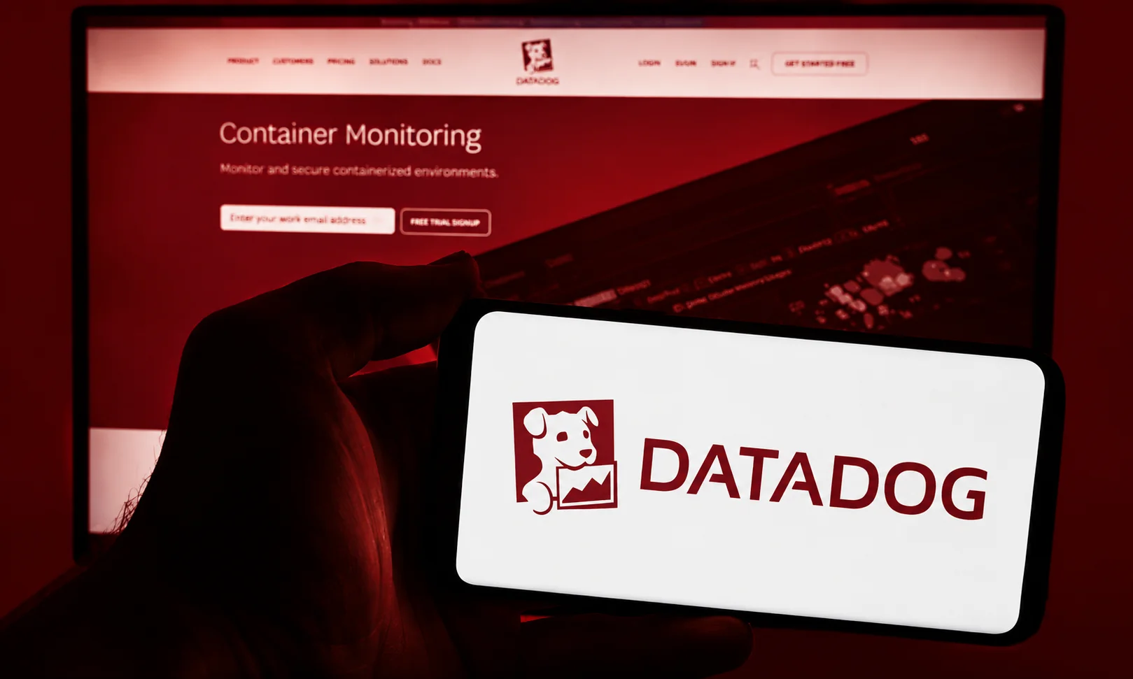 A person holding a mobile phone showing the Datadog interface with a container monitoring dashboard on a desktop screen behind it.