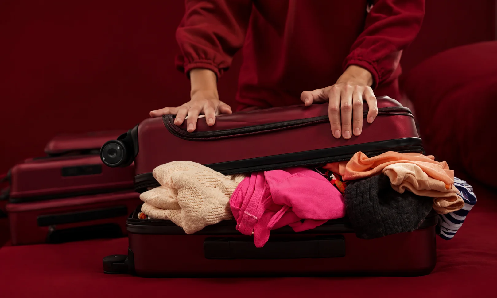 Hands arranging items inside a suitcase set against a dark red surface.