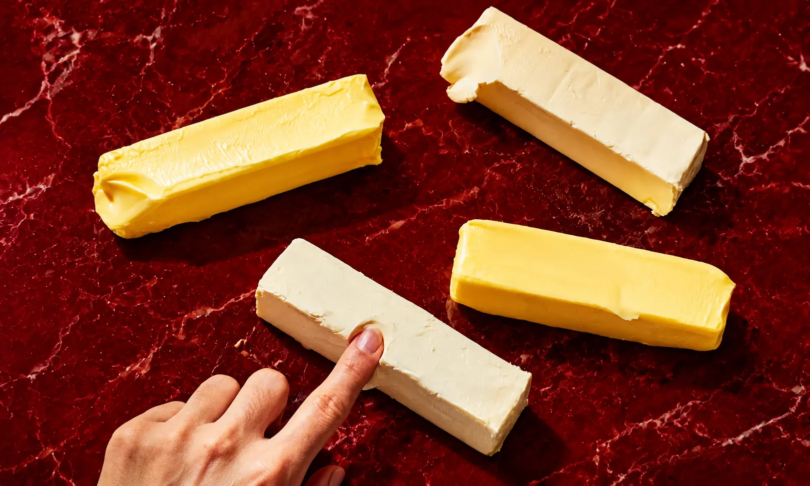 A person using a knife to slice a block of softened butter on a plate against a deep red background.
