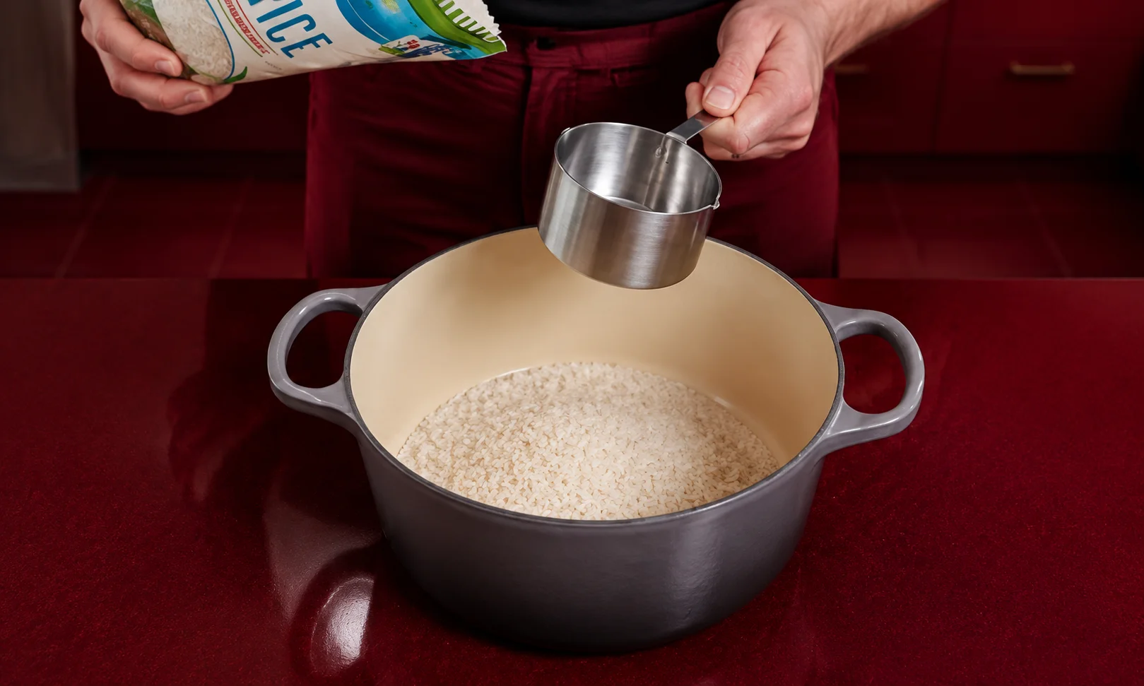 Hands measuring rice into a cooking pot displayed against a rich red studio backdrop.