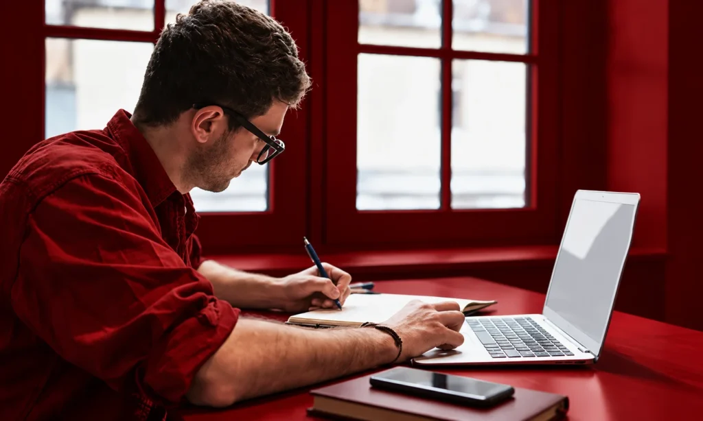 | How Brands Are Rewriting Content for Answer Engines A man in a red shirt and glasses writes in a notebook beside an open laptop at a red desk, representing careful content rewriting for AEO on an existing page.